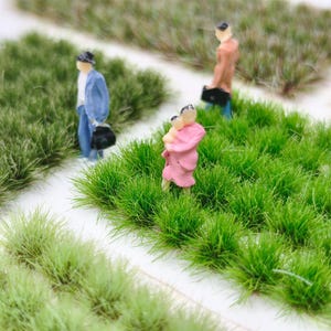 May include: Miniature figurines of people walking through a grassy area. One figure wears a blue suit and hat, another a brown coat, and a third carries a child in a pink coat. The grass is a vibrant green, and the scene is set on a white surface.