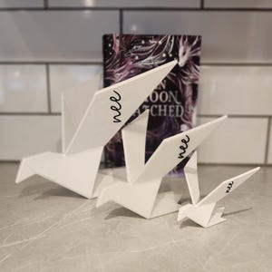 May include: Three white origami-style bird sculptures of varying sizes, each featuring the word "nee" in black cursive script on one wing. The birds are arranged in a row, with the largest on the left and the smallest on the right, positioned in front of a book.