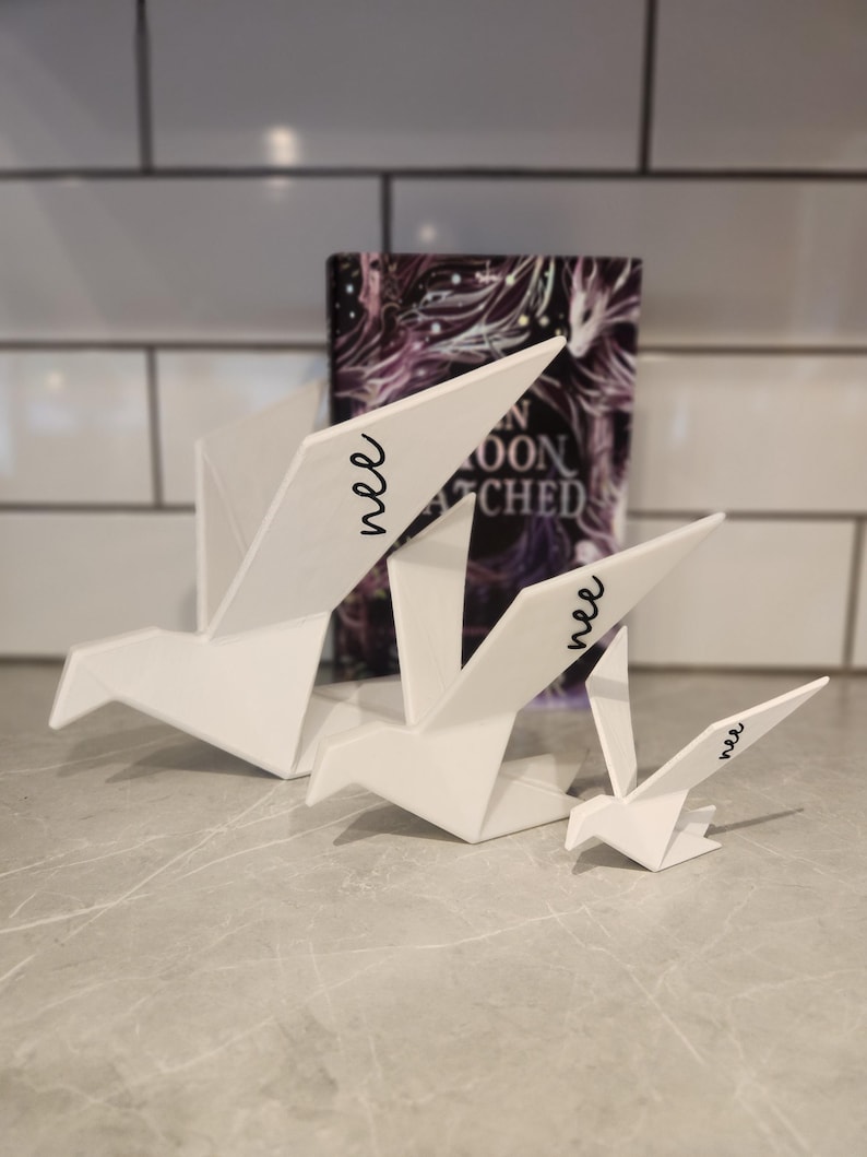 May include: Three white origami-style bird sculptures of varying sizes, each featuring the word "nee" in black cursive script on one wing. The birds are arranged in a row, with the largest on the left and the smallest on the right, positioned in front of a book.