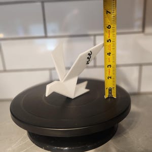 May include: A white origami-style bird figurine with the word "nee" written in black script on its wing. A yellow measuring tape is positioned next to the bird, indicating a height of approximately 6.5 inches from the base to the wing tip.