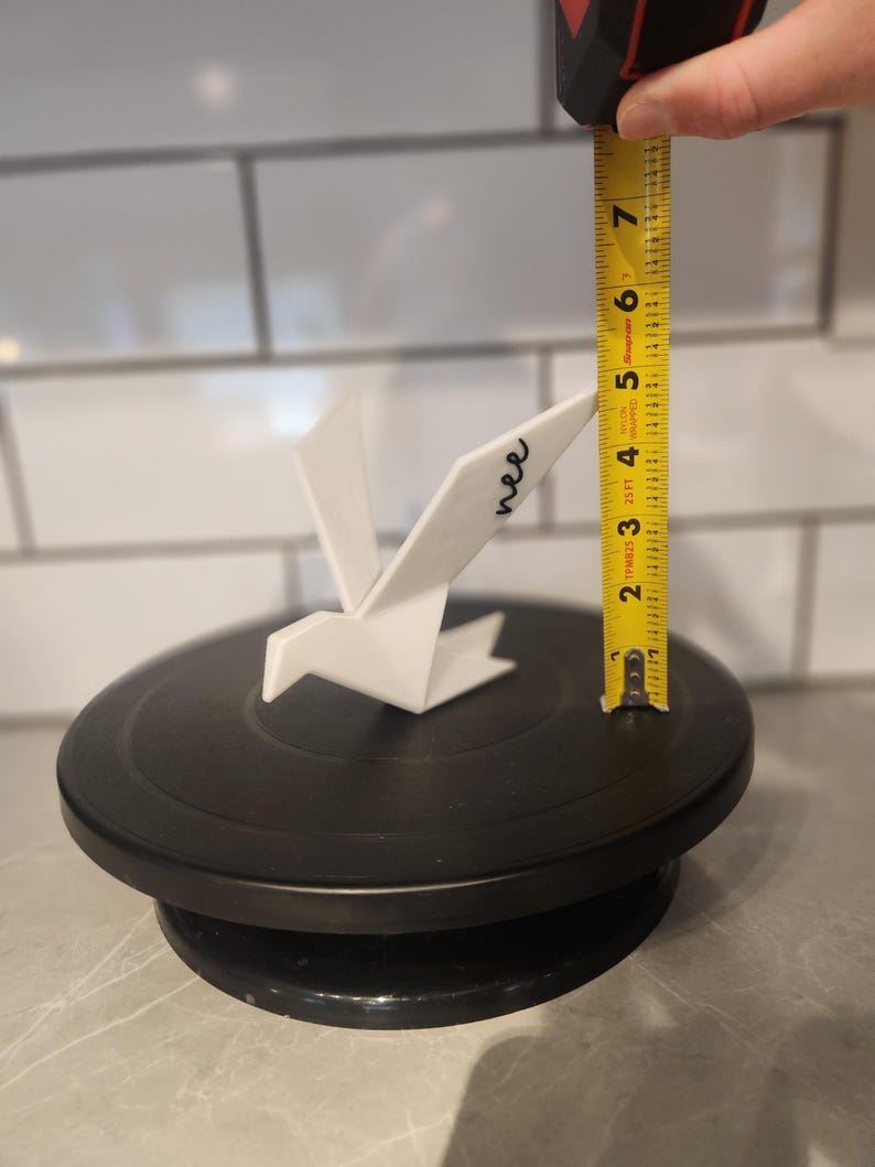 May include: A white origami-style bird figurine with the word "nee" written in black script on its wing. A yellow measuring tape is positioned next to the bird, indicating a height of approximately 6.5 inches from the base to the wing tip.