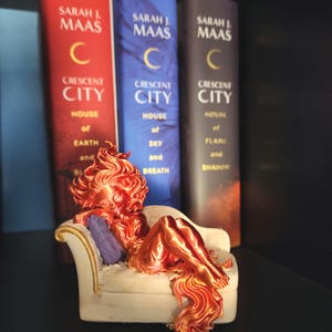 May include: A copper-colored figurine of a reclining figure on a small white couch, with three books in the background. The books are titled "Crescent City" by Sarah J. Maas. The books are red, blue, and dark grey.