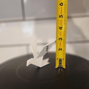 May include: A white, bird-shaped ornament with the word "nee" written in black script on its wing. A yellow measuring tape stands vertically next to the ornament, indicating a height of approximately 2.25 inches from the black surface base.