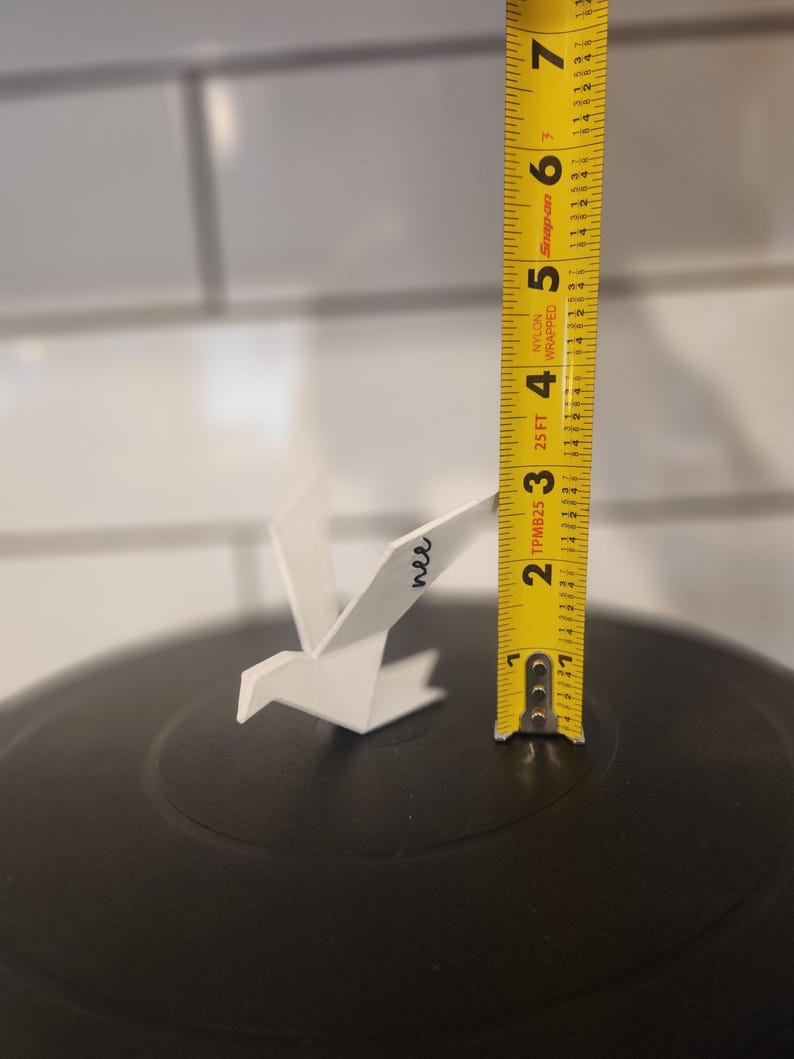 May include: A white, bird-shaped ornament with the word "nee" written in black script on its wing. A yellow measuring tape stands vertically next to the ornament, indicating a height of approximately 2.25 inches from the black surface base.