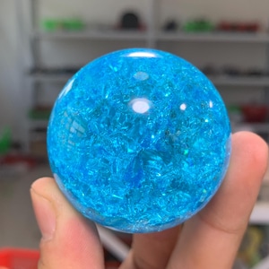 May include: A spherical object made of clear material containing a dense cluster of bright blue, jagged, crystalline fragments. The sphere has a smooth, polished outer surface that reflects light, creating a bright white highlight on the top.