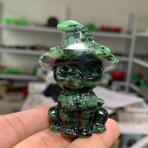 May include: A small, carved stone figurine of a cat wearing a pointed hat. The sculpture is made of a polished green and black mottled mineral, featuring a smooth, glossy surface that highlights the natural dark inclusions within the stone material.