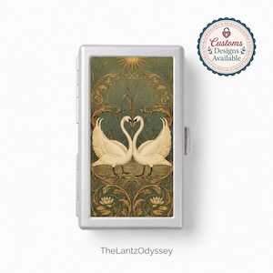 May include: A rectangular silver metal case featuring a central illustration of two white swans facing each other with necks forming a heart shape, set against a dark green background with gold filigree, water lilies, and a sun, with the text TheLantzOdyssey.