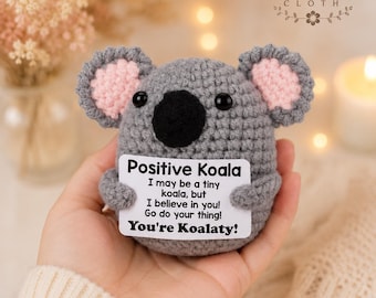 May include: A small gray crocheted koala toy with pink inner ears and black eyes. The koala holds a white rectangular sign that reads: Positive Koala, I may be a tiny koala, but I believe in you! Go do your thing! You're Koalaty!