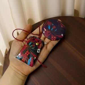 May include: A rectangular fabric omamori charm and a circular pin button, both featuring a dark-themed illustration of a hooded figure in black, red, and blue with a white cross symbol, accompanied by a thin maroon cord for hanging the charm.