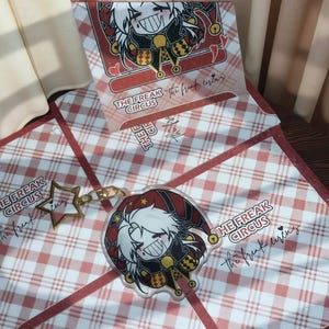 Puede incluir: Un llavero acrílico transparente y una tarjeta a juego con un personaje de dibujos animados con cabello blanco y una sonrisa traviesa. El llavero tiene un dije de estrella dorada. El texto "THE FREAK CIRCUS" es visible.