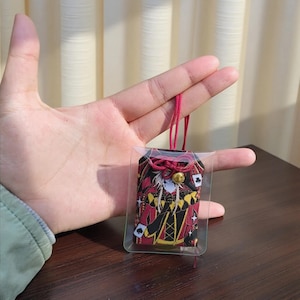 Pu&ograve; includere: Un portafortuna a tema carte da gioco in una custodia di plastica trasparente, con colori rosso, nero e oro. Il ciondolo presenta un design di personaggi con simboli di carte da gioco. &Egrave; attaccato un cordino rosso con una campana d'oro.