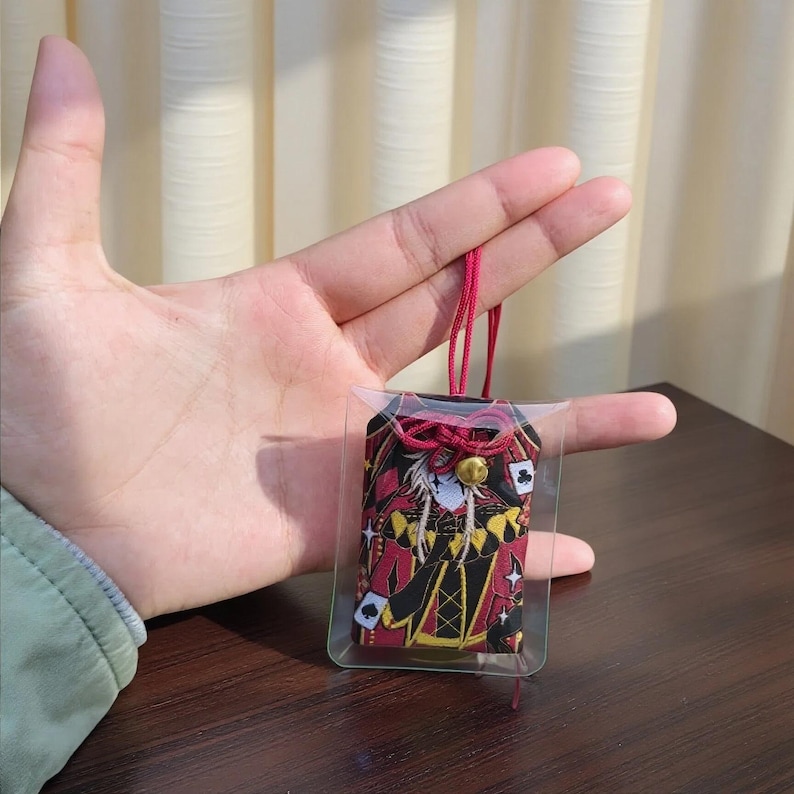 Pu&ograve; includere: Un portafortuna a tema carte da gioco in una custodia di plastica trasparente, con colori rosso, nero e oro. Il ciondolo presenta un design di personaggi con simboli di carte da gioco. &Egrave; attaccato un cordino rosso con una campana d'oro.