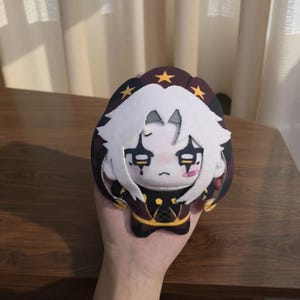 May include: A plush doll with a white face, white hair, and a black and red outfit. The doll has a sad expression with black markings around the eyes and a small pink tongue. The doll is held in a hand.