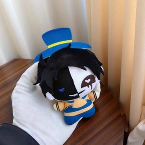 Könnte beinhalten: Plüschpuppe mit schwarz-weißem Gesicht, schwarzen Haaren und einem blauen Hut mit gelbem Band. Die Puppe trägt ein blau-gelb gestreiftes Outfit. Die Puppe ist ein Sammlerstück.
