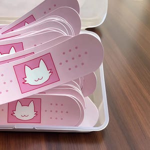 Könnte beinhalten: Rosa Pflaster mit einem weißen Katzen-Gesicht-Design in einem transparenten Kunststoffbehälter. Die Pflaster haben einen rosa Hintergrund mit einem weißen Katzen-Gesicht in einem rosa Quadrat. Die Pflaster sind oval geformt mit kleinen rosa Punkten.