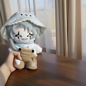 May include: A plush doll with light blue hair and a matching hat, wearing a white outfit and a light blue scarf. A small brown frog-shaped keychain with a star and bear design is held in front of the doll.