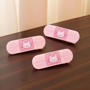 Puede incluir: Tres pinzas para el pelo rosas con forma de vendaje con un diseño de cara de gato. Cada pinza tiene un fondo rosa claro con una cara de gato blanca en un cuadrado rosa. Las pinzas están sobre una superficie de madera marrón oscuro.