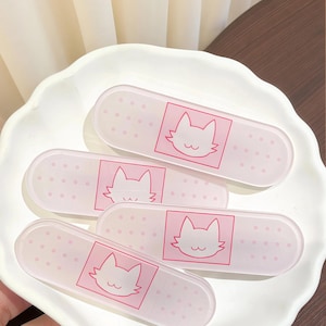 Könnte beinhalten: Vier rosa Pflaster mit einem Katzen-Gesicht-Design. Jedes Pflaster hat ein rosa Katzen-Gesicht in einem rosa Quadrat mit kleinen rosa Punkten. Die Pflaster liegen auf einem weißen, blattförmigen Teller.