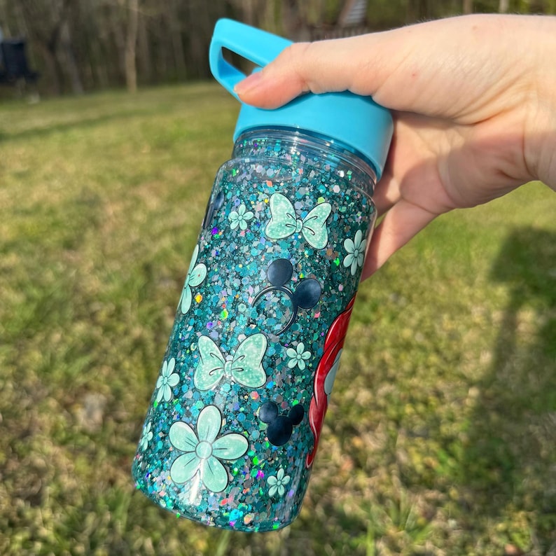 May include: A clear water bottle filled with teal glitter and decorated with light blue bows, flowers, and Mickey Mouse silhouettes. The bottle has a light blue lid and handle. The bottle is held by a hand against a blurred green background.
