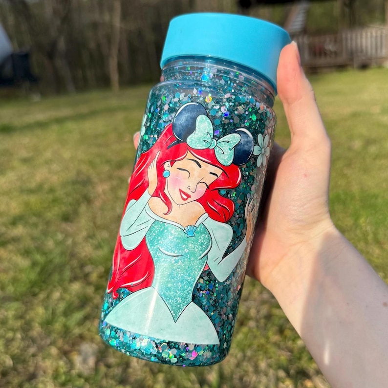 May include: A clear water bottle filled with glitter, featuring a light blue lid. The bottle is decorated with a cartoon image of Ariel from The Little Mermaid, with red hair and a teal dress. The bottle is held in a hand.