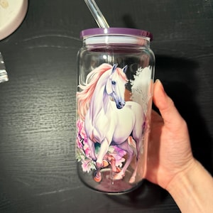 May include: A clear glass tumbler with a purple lid and straw. The tumbler features a watercolour design of a white horse with a pink mane and floral accents. The tumbler is designed for cold beverages.