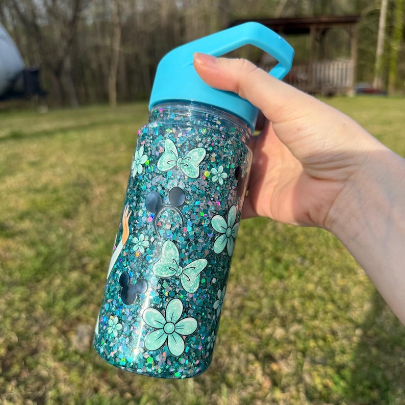 May include: A teal glitter water bottle with a light blue handle and lid. The bottle is decorated with light blue butterflies, flowers, and Mickey Mouse silhouettes. The bottle is held in a person's hand.