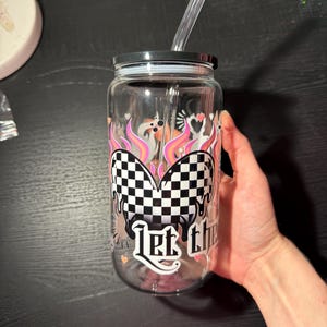May include: A clear glass tumbler with a black lid and a clear straw. The tumbler has a design featuring a checkered heart, flames, and the words "Let the". The design incorporates pink, white, and black colours.