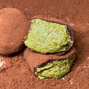May include: Close-up of chocolate truffles dusted with cocoa powder. One truffle is cut open, revealing a green pistachio filling. The truffles are arranged on a bed of cocoa powder, creating a rich, textured appearance.