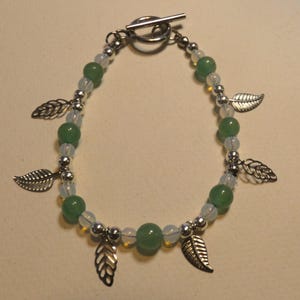 Könnte beinhalten: Ein silberfarbenes Armband mit grünen und weißen Perlen und Blattanhängern. Das Armband hat einen Knebelverschluss und ist mit kleinen silbernen Perlen aufgereiht. Die grünen Perlen sind mit weißen Perlen und silbernen Blattanhängern durchsetzt.