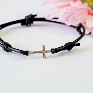 May include: A black leather bracelet with a silver cross charm. The bracelet is tied with a knot on each end.