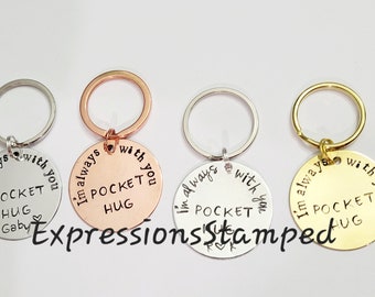 May include: Four round keychains in silver, rose gold, silver, and gold. Each keychain has a ring and a circular charm with the words "I'm always with you" and "Pocket Hug" engraved. The text "Expressions Stamped" is also visible.