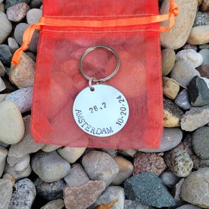 May include: A silver keychain with the text "AMSTERDAM 26.2 10-20-22" on a round tag. The keychain is in a red drawstring bag and on a bed of gray rocks.
