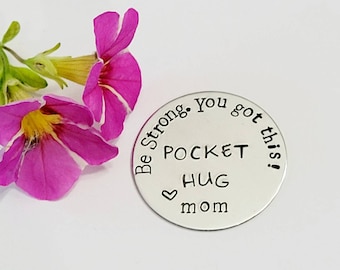 May include: Silver round charm with the text "Be Strong. You got this!"  "Pocket Hug" and a heart symbol with the word "mom" below.