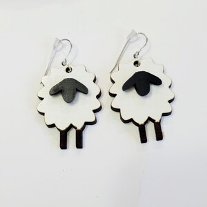 May include: A pair of white wooden sheep earrings with black faces and brown legs. The earrings are hanging from silver hooks.