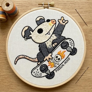 Puede incluir: Arte de aro bordado con un dibujo de zarigüeya en monopatín. La zarigüeya es negra, blanca y marrón, con un monopatín que tiene un diseño de calavera y huesos cruzados con llamas. El texto "POSSUM.MOOD" está en el monopatín.