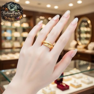 May include: A gold-colored ring with a double-band design is displayed on a hand. The ring is smooth and shiny, reflecting the light. The background is a jewelry store setting with display cases and the Hollow Finch logo.