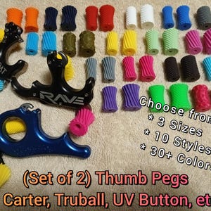 May include: A collection of brightly coloured thumb pegs for archery release aids, with three release aids. The pegs are available in over 30 colours. Text on the image reads "Choose from: 3 Sizes, 10 Styles, 30+ Colours."