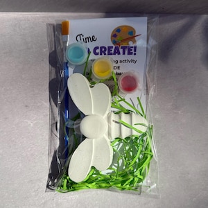 May include: A clear plastic bag contains a craft kit with a white bunny-shaped figure, paint pots in blue, yellow, and red, a paintbrush, and green paper grass. The kit includes the text "Time to CREATE!"