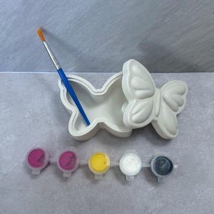 May include: A white butterfly-shaped ceramic box with a lid, a blue paintbrush, and a set of paint pots. The paint colors include pink, yellow, white, and gray. The art supplies are arranged on a gray surface.