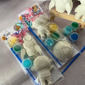May include: Paint-your-own space-themed craft kits. Each kit includes a rocket ship, astronaut, and planet figurine, along with paint pots and a paintbrush. The kits are packaged in clear plastic bags with colorful confetti.