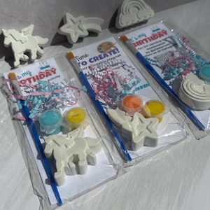 May include: DIY paintable magnet kits for a birthday activity. Each kit includes a paint brush, paint pots, and a plaster figure in the shape of a unicorn, star, or rainbow. The kits are packaged in clear bags with colorful confetti.