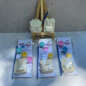 May include: A craft kit featuring backpack-shaped figurines, paint pots in blue, pink, and yellow, and paintbrushes. The packaging reads "CREATE & ENJOY" and "your-own master kit made with care". The kit includes instructions for painting the figurines.