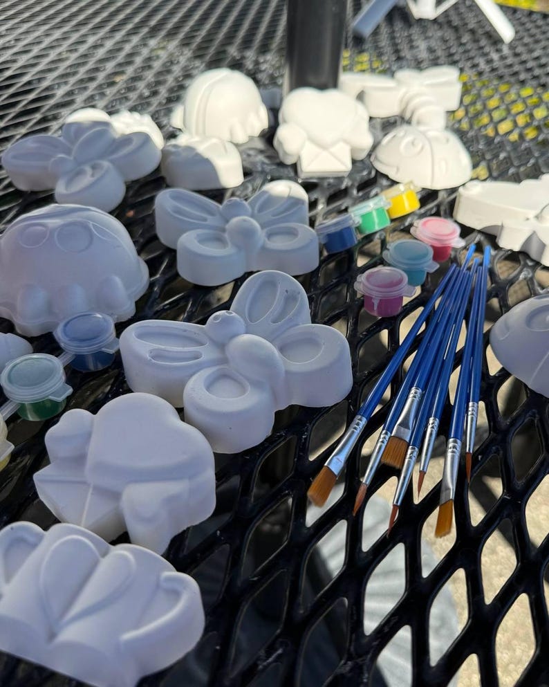 May include: A collection of unpainted ceramic figurines in various shapes, including butterflies, hearts, and ladybugs. Small paint pots with assorted colors and several blue-handled paintbrushes are also present, suggesting a DIY craft kit.