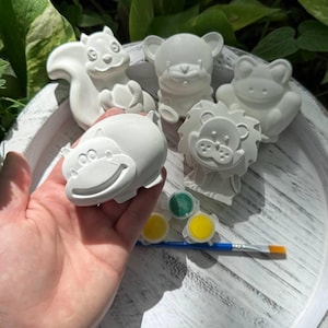 May include: A collection of unpainted ceramic animal figurines, including a squirrel, bear, frog, lion, and hippo. The set includes paint pots in green and yellow, and a paintbrush. The figurines are on a white wooden tray.