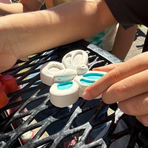 May include: A white, butterfly-shaped mold with four sections, each containing a pool of bright blue liquid. The mold is held by a person's hand, with the background showing other people and a black metal surface.