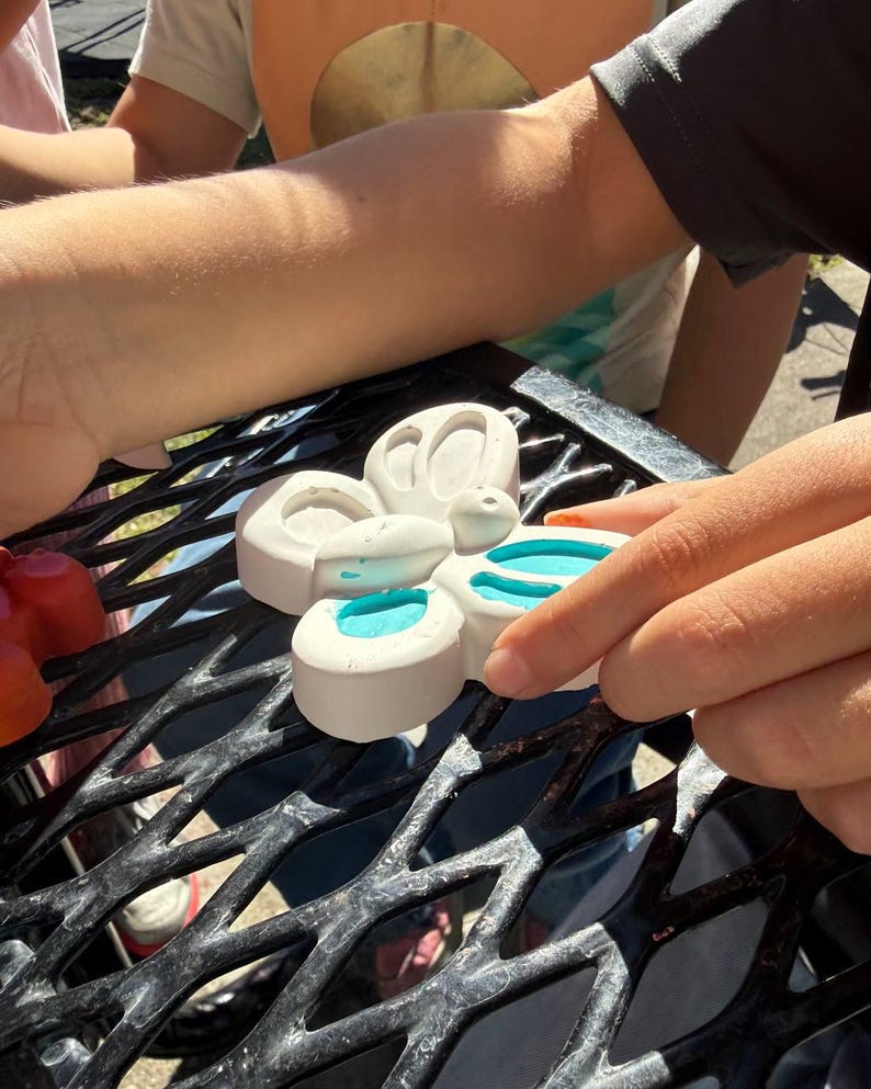 May include: A white, butterfly-shaped mold with four sections, each containing a pool of bright blue liquid. The mold is held by a person's hand, with the background showing other people and a black metal surface.