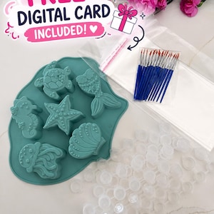 May include: A teal silicone mold with sea creature shapes, paintbrushes, paint pots, and a clear bag. The text "FREE DIGITAL CARD INCLUDED!" is displayed in pink and white. Pink flowers are in the background.