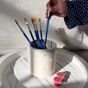 May include: A white cylindrical container with heart-shaped cutouts holds several paintbrushes with blue handles and yellow bristles. Two small red paint pots sit on a white, distressed wooden tray. A hand with a blue polka-dot sleeve holds a paintbrush.