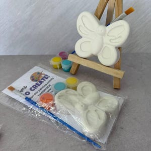 May include: A DIY painting kit featuring a butterfly-shaped ceramic figure, paint pots in various colors, and paintbrushes. The kit includes the text "time TO CREATE!" and is displayed on a small wooden easel.