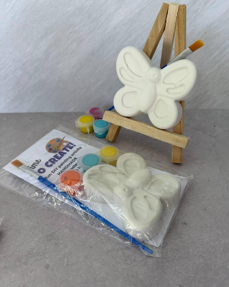 May include: A DIY painting kit featuring a butterfly-shaped ceramic figure, paint pots in various colors, and paintbrushes. The kit includes the text "time TO CREATE!" and is displayed on a small wooden easel.